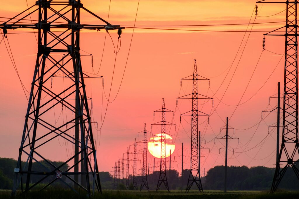 A line of high-voltage electricity pylons receding into the distance, silhouetted against the setting sun in an orange sky