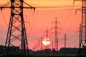 A line of high-voltage electricity pylons receding into the distance, silhouetted against the setting sun in an orange sky
