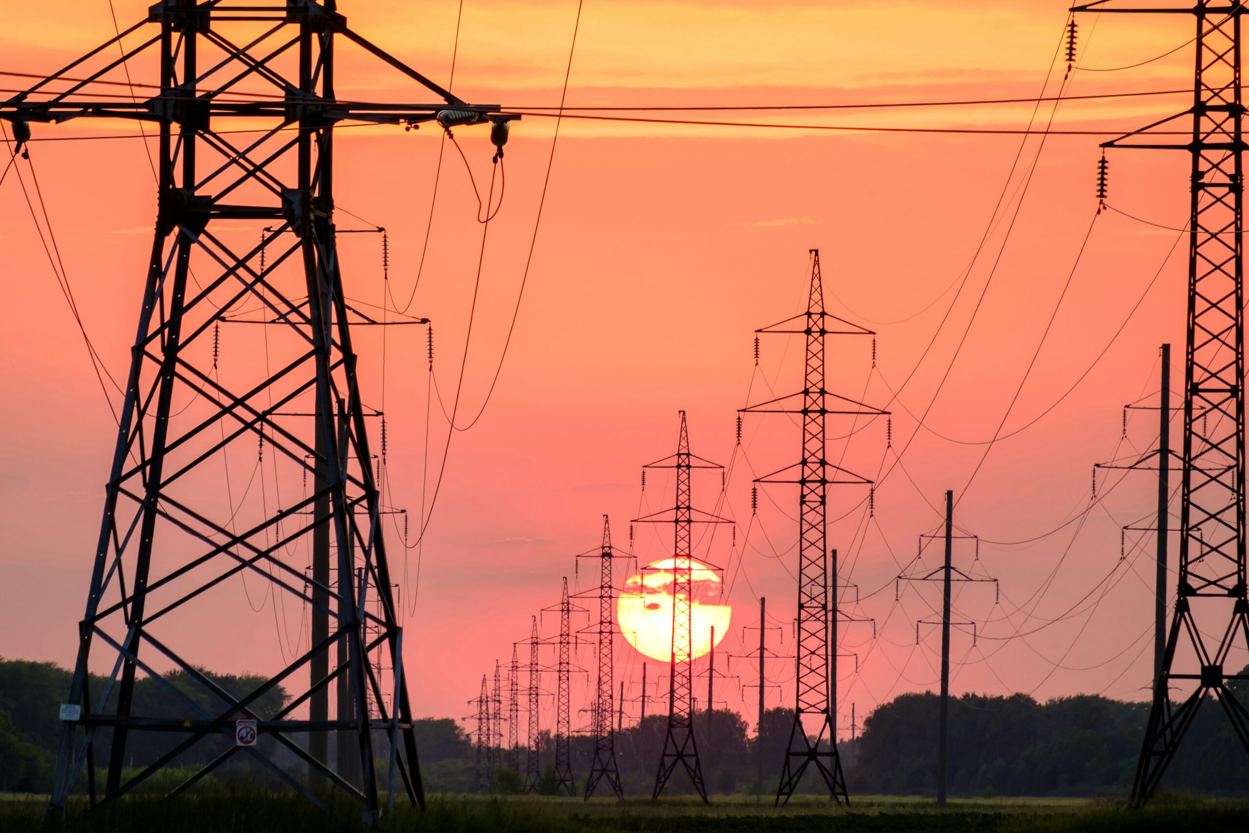 A line of high-voltage electricity pylons receding into the distance, silhouetted against the setting sun in an orange sky
